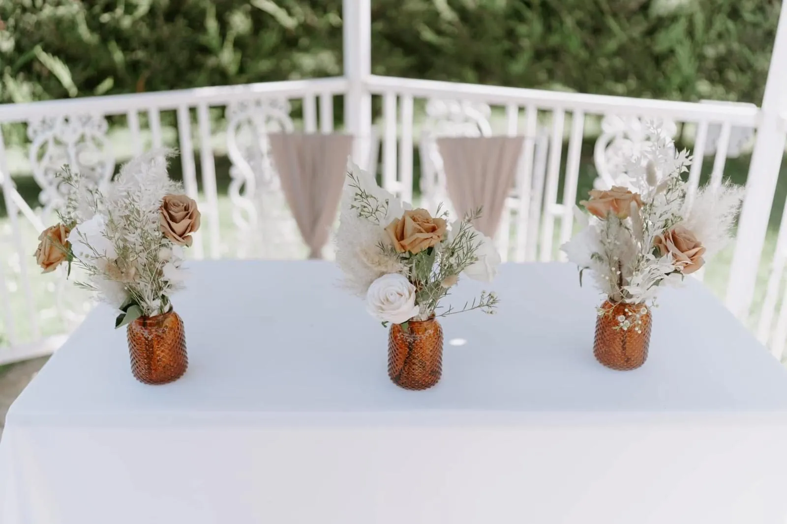 Three amber glass vases holding toffee roses, white blooms and pampas grass on a white-clothed top table